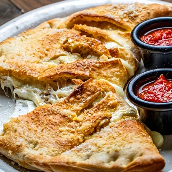 Golden-brown calzone, sliced open to reveal melted cheese, served with two cups of marinara sauce.